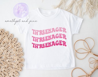 May include: White t-shirt with a pink and white ombre graphic that says "Three-nager" three times.
