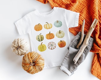 May include: White t-shirt featuring a watercolor design of various pumpkins in shades of orange, green, yellow, and white. The shirt is paired with gray pants, decorative twine balls, and an orange fabric. A fall-themed children's apparel.