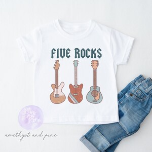 May include: White t-shirt with the text "FIVE ROCKS" above three illustrated guitars in shades of brown, red, and blue. A pair of blue jeans is folded next to the shirt. The shirt has a round neck and short sleeves.