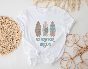 May include: White t-shirt with a graphic of three surfboards with different patterns and the text "Surfer Mom" in a retro font.