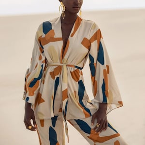 Può includere: Una donna indossa un kimono di seta beige, arancione e blu con un motivo, con un paio di pantaloni abbinati. Il kimono ha una cintura a nodo e maniche larghe. La donna indossa orecchini e anelli d'oro.