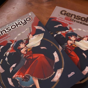 Puede incluir: Dos libros de tapa blanda titulados Chroniques de Gensokyo y Gensokyo Chronicles. Las portadas presentan un fondo azul oscuro con una ilustración central de un personaje con traje rojo sosteniendo amuletos de papel blanco, con el texto Azulia.