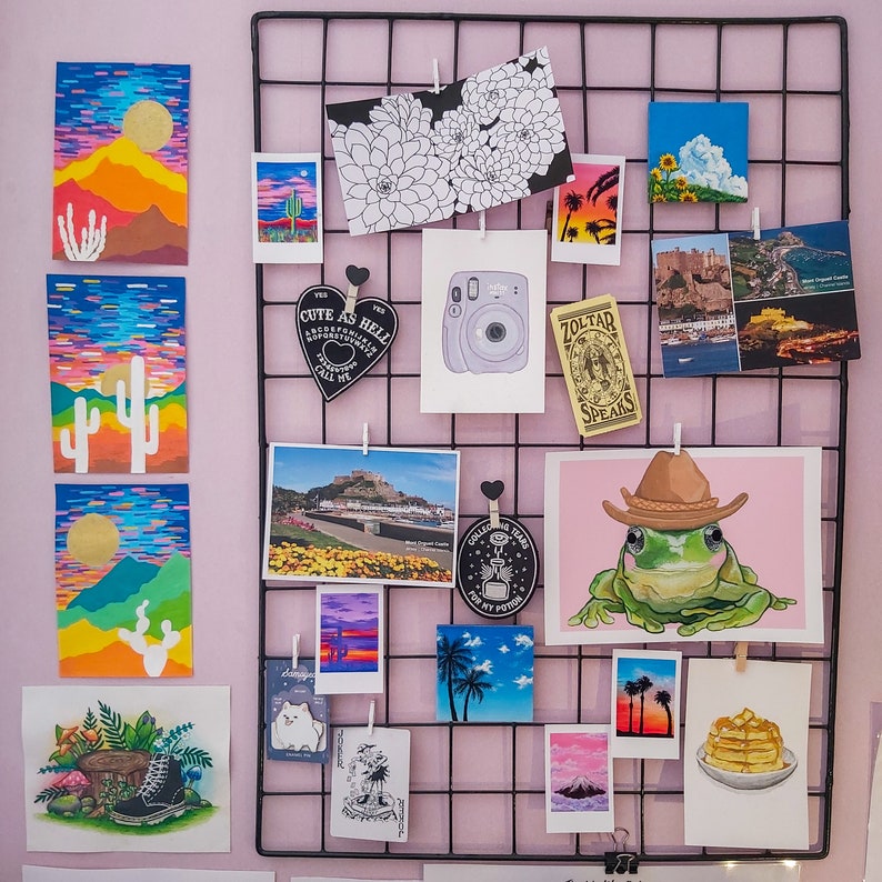 Peut inclure: Une grille en fil de fer noir avec diverses cartes et images color&eacute;es &eacute;pingl&eacute;es dessus. Les cartes pr&eacute;sentent une vari&eacute;t&eacute; de motifs, notamment une grenouille, un cactus, une fleur, un personnage de dessin anim&eacute; et un paysage urbain. Les textes "Cute as Hell" et "Zoltar Speaks" sont visibles sur deux des cartes.