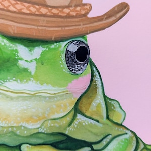 Peut inclure: Une grenouille verte portant un chapeau de cowboy brun. La grenouille a un gros &oelig;il noir et un fard &agrave; joues rose. L'arri&egrave;re-plan est rose clair.