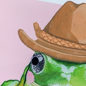 Peut inclure: Gros plan sur une grenouille verte portant un chapeau de cowboy brun. L'&oelig;il de la grenouille est visible et le chapeau a une bande tiss&eacute;e.