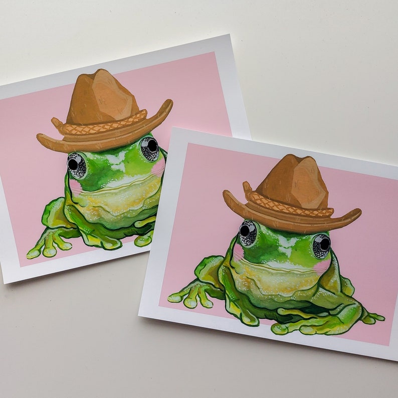 Peut inclure: Deux grenouilles vertes portant des chapeaux de cowboy bruns sur un fond rose clair. Les grenouilles sont face avant, la bouche ferm&eacute;e. Les chapeaux ont un bandeau brun avec un motif tress&eacute;.