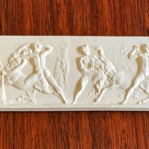 May include: A rectangular off-white ceramic plaque featuring a raised relief design of classical figures, including humans and centaurs in dynamic poses. The surface is matte and monochromatic, showcasing intricate sculptural details of the figures.