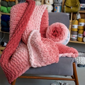 May include: A plush, hand-knitted blanket in shades of pink and red is draped over a gray chair. A matching ball of yarn sits beside the blanket. The background features shelves filled with various colors of yarn.