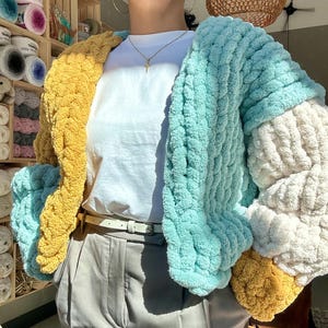 May include: A chunky knit cardigan in a patchwork of colors: yellow, light blue, and cream. The open-front sweater is made of thick, soft yarn. The sleeves are a mix of colors, and the garment is worn over a white t-shirt and gray pants.