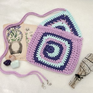 May include: A crocheted lavender and light blue purse with a geometric pattern. The purse has a long strap and is decorated with two silver crescent moon charms.