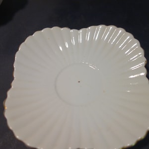 May include: White saucer with a scalloped edge and a center depression for a cup. The saucer has a subtle sheen and a delicate, feminine design.