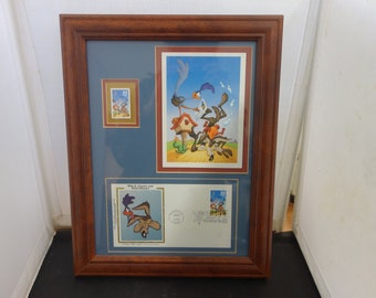 May include: Framed artwork featuring Looney Tunes characters Wile E. Coyote and Road Runner. The frame holds a large print, a smaller stamp, and a first-day cover, all with cartoon imagery. The frame is brown, and the background is blue.