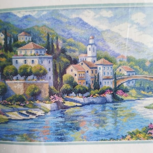 Puede incluir: Paisaje de punto de cruz que representa una escena de pueblo europeo. La imagen presenta edificios con tejados de tejas rojas, un puente de piedra y un río que refleja el cielo. La paleta de colores incluye azules, verdes y tonos tierra, creando un ambiente sereno.