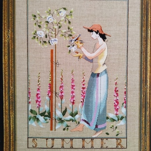 May include: Cross-stitch artwork in a gold frame depicts a woman in a hat tending a flowering tree. The scene includes pink and green flowers, with the word "SUMMER" stitched at the bottom. The woman wears a yellow top and blue skirt.