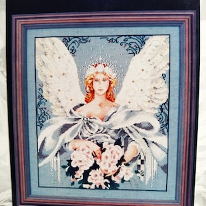 Op de afbeelding: Ingelijst kruissteekkunstwerk met een engel met witte vleugels, een halo en een vloeiende jurk. De engel houdt een boeket roze rozen vast. Het kunstwerk is ingelijst in een houten lijst met een blauwe en bruine rand.