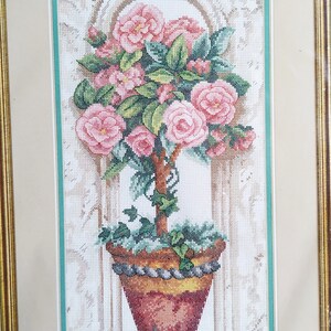 Puede incluir: Un bordado en punto de cruz enmarcado que representa un topiario de rosas en maceta con flores rosas y hojas verdes. La planta está en una maceta marrón y amarilla con un borde gris decorativo, sobre un fondo claro con un arco arquitectónico.