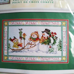 May include: A Dimensions counted cross stitch kit titled Family Christmas by Donna Race. The package features a winter scene with three snowmen, a sled, birdhouses, and a small dog. The text Merry Christmas appears in red on the border.