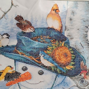 May include: A cross-stitch pattern featuring a snowman wearing a blue hat decorated with a sunflower. Several birds, including a cardinal, blue jay, and goldfinch, surround the hat. A bright orange carrot nose is visible on the snowman's face.
