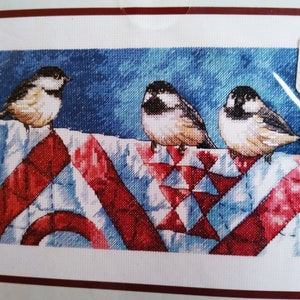 May include: A cross-stitch embroidery pattern featuring three small birds with black heads and white cheeks perched on a red, white, and blue geometric quilt hanging on a clothesline against a dark blue textured background.