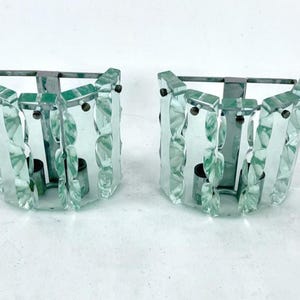 Op de afbeelding: Twee bijpassende wandkandelaars met een helder, facetgeslepen glazen ontwerp en een metalen frame. De wandkandelaars hebben een lichtgroene tint en zijn ontworpen om kaarsen te bevatten. De algehele stijl is vintage en decoratief.