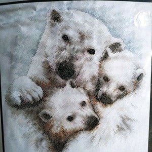 Puede incluir: Un patrón de bordado de punto de cruz que muestra un oso polar blanco grande con dos oseznos más pequeños. El diseño utiliza tonos de hilo blanco, crema y marrón para crear la textura del pelaje sobre un fondo de cuadrícula azul claro y blanco.