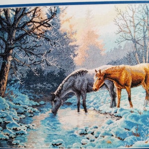 Puede incluir: Un patrón de bordado en punto de cruz que representa un caballo gris y un caballo marrón bebiendo de un arroyo en un bosque nevado. La escena presenta árboles desnudos, nieve con tonos azules y luz suave, creada con puntadas detalladas.