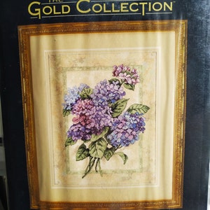 Könnte beinhalten: Ein Dimensions The Gold Collection Zählstickpackung-Set mit dem Titel Elegant Hydrangeas. Die Verpackung zeigt ein florales Design aus violetten und blauen Hortensienblüten mit grünen Blättern auf elfenbeinfarbenem Stoff, 28 cm mal 36 cm groß.