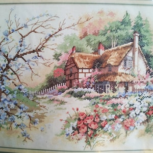 May include: A cross-stitch embroidery depicting a cottage with a thatched roof, timber-framed walls, and two chimneys. The scene includes a large tree with blue blossoms on the left, a white picket fence, and a foreground filled with red and white flowers.