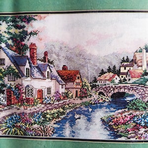 May include: A cross-stitch embroidery depicting a rural landscape with a stone cottage, a stone bridge over a flowing stream, and two white swans. The scene features colourful flower gardens, green trees, and a mountainous backdrop in soft pastel tones.