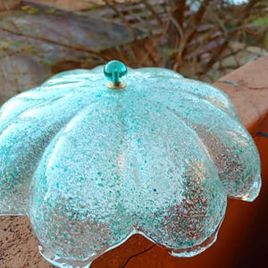 May include: A clear, turquoise glass decorative bowl with a flower-like design. The bowl features a speckled pattern and a small, round, turquoise knob on top. The knob is attached to a gold-coloured base.