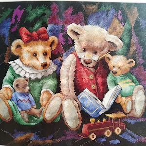 May include: A cross-stitch embroidery depicting a family of four teddy bears. Two adult bears sit with two smaller bears, one wearing a blue jumper and one in a green jumper. The larger bear wears glasses and a red waistcoat, reading a blue book near a toy train.