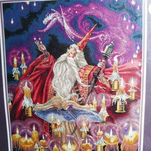 May include: A cross-stitch embroidery depicting a wizard with a long white beard, wearing a red robe and pointed hat, holding a staff. The wizard stands before an open book surrounded by numerous lit candles against a dark purple background.