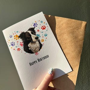 May include: A white greeting card with a black and white dog illustration and colorful paw prints. The words "HAPPY BIRTHDAY" are printed below the dog. The card is paired with a brown envelope.