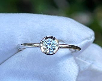 May include: A silver ring with a round, clear gemstone set in a bezel. The ring is displayed against a white background, with a blurred green backdrop.