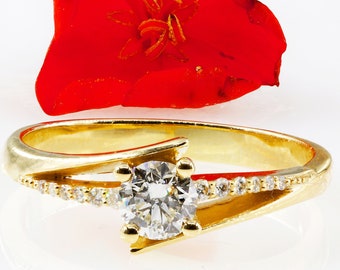 Op de afbeelding: Een gouden ring met een grote diamant en kleinere diamanten die in een band zijn gezet. De ring ligt op een witte achtergrond met een rode bloem.