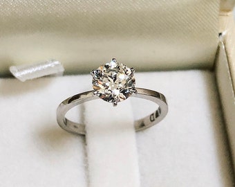 May include: A silver engagement ring with a round, clear diamond in a six-prong setting. The ring is in a white box.