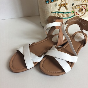 May include: A pair of white leather sandals with brown straps and soles. The sandals have a criss-cross design on the top of the foot.