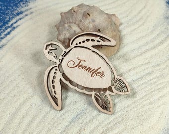 Puede incluir: Una tortuga marina de madera con el nombre "Jennifer" grabado. La tortuga, de color marrón claro, presenta intrincados detalles calados. Descansa sobre arena blanca, con una concha en el fondo.