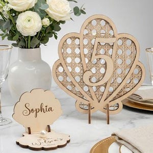 Könnte beinhalten: Eine Tischdekoration mit einer Vase weißer Rosen, einer tischförmigen Tischkarte in Muschelform mit der Nummer 5 und einer Tischkarte aus Holz mit dem Namen "Sophia". Goldgeränderte Teller, Gläser und Besteck vervollständigen die elegante Anordnung.