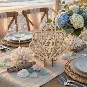 Op de afbeelding: Een tafelsetting met strandthema met een houten tafelnummer vier in de vorm van een schelp. De tafel is gedekt met borden, glazen en een bloemstuk met blauwe en witte hortensia's. Schelpen en koraal zijn over de tafel verspreid.