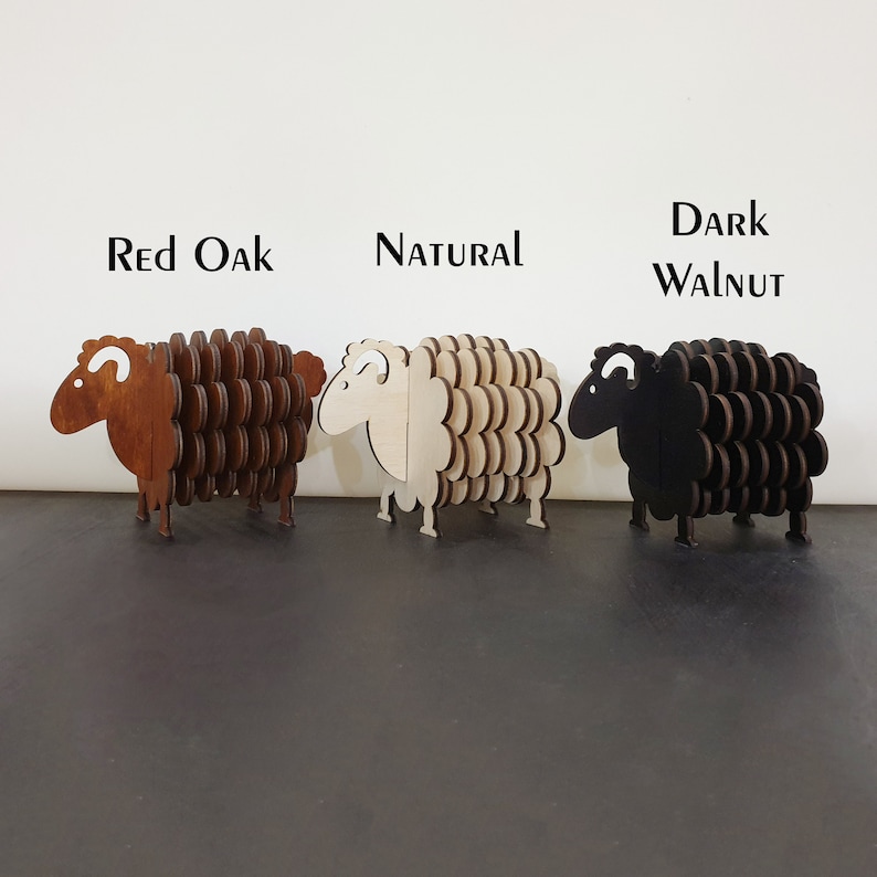 May include: Three wooden sheep coasters in different wood stains: red oak, natural, and dark walnut. The coasters are shaped like sheep with a curly tail and a small head.