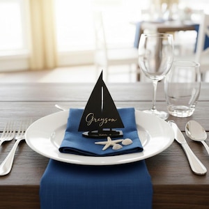 Puede incluir: Una mesa de tem&aacute;tica n&aacute;utica con un tarjetero de barco negro que dice "Greyson" en escritura dorada. El montaje incluye una servilleta azul, un plato blanco, cubiertos y conchas decorativas.