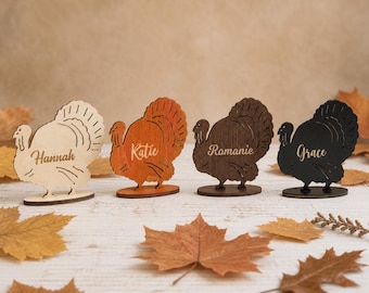 Puede incluir: Cuatro tarjetas de sitio de madera con forma de pavo sobre bases ovaladas. Cada pavo tiene un tono de madera diferente, de claro a oscuro, y presenta un nombre grabado con láser: Hannah, Katie, Romanie y Grace, en letra cursiva.