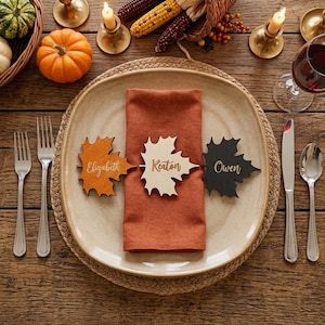 May include: Three maple leaf-shaped wooden place cards in orange, cream, and black, each engraved with the names Elizabeth, Keaton, and Owen, resting on a folded rust-coloured linen napkin atop a ceramic plate with silver cutlery on the sides.