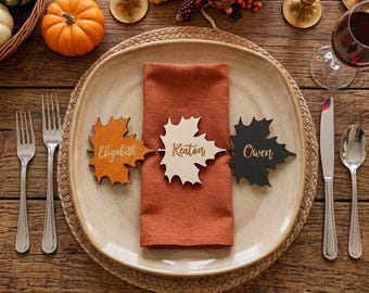 May include: Three maple leaf-shaped wooden place cards in orange, cream, and black, each engraved with the names Elizabeth, Keaton, and Owen, resting on a folded rust-colored linen napkin atop a ceramic plate with silver cutlery on the sides.