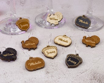 Puede incluir: Un conjunto de nueve encantos para copas de vino con temática de Acción de Gracias. Los encantos están hechos de madera y presentan diferentes diseños, que incluyen pavos, bellotas y calabazas. Cada encanto está personalizado con un nombre, incluidos "Jenny", "William" y "Michelle".