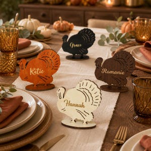Puede incluir: Cuatro tarjetas de sitio de madera con forma de pavo sobre una mesa. Cada pavo es de un color distinto: naranja, negro, marrón oscuro y madera clara natural. Los nombres Katie, Grace, Romanie y Hannah están impresos en letras doradas.