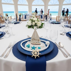 Puede incluir: Decoraci&oacute;n de mesa de tem&aacute;tica n&aacute;utica con mantel blanco, platos azules y un velero decorativo. El conjunto incluye un tim&oacute;n dorado, estrellas de mar y un adorno de cuerda. El nombre "Harry" est&aacute; escrito en el velero.