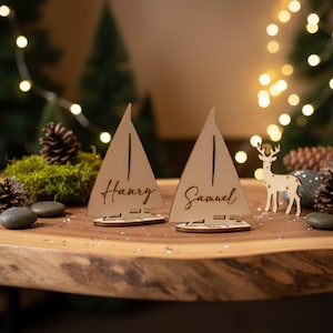 Puede incluir: Dos portatarjetas de madera con forma de velero con los nombres "Harry" y "Samuel" grabados. Las tarjetas est&aacute;n sobre una mesa de madera, con un peque&ntilde;o ciervo de madera y pi&ntilde;as de fondo.
