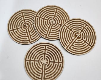 May include: Four circular wooden coasters featuring intricate laser-engraved labyrinth patterns. Each coaster displays a unique central symbol, including a flame, a cross, and a floral motif, rendered in dark brown lines against a light wood.